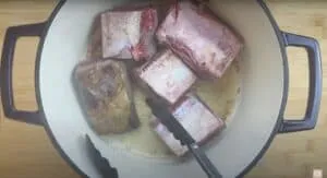 Browning beef short ribs in a Dutch oven with tongs on a wooden cutting board background.